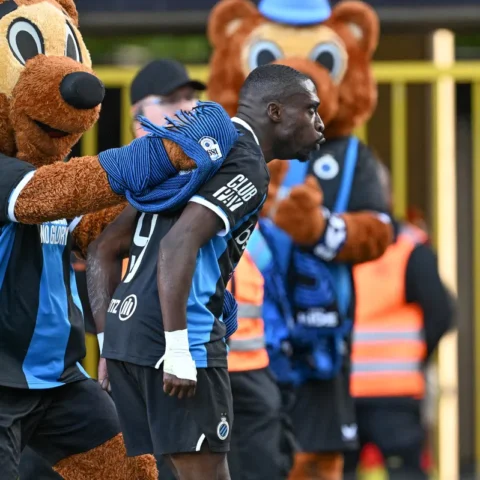 Een Club Brugge-speler loopt met een mascotte op de achtergrond bij het stadion.