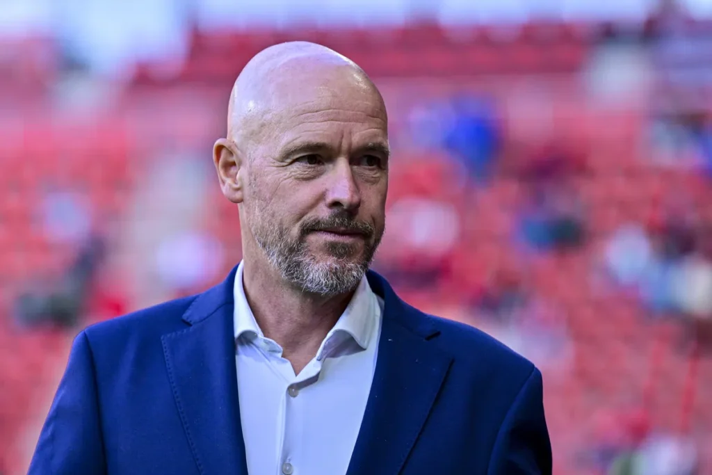 Erik ten Hag in een donkerblauw colbert voor een onscherpe stadionachtergrond.