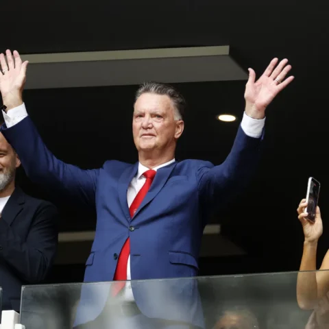 Louis van Gaal staat in een skybox met beide handen omhoog terwijl omstanders applaudisseren en filmen.