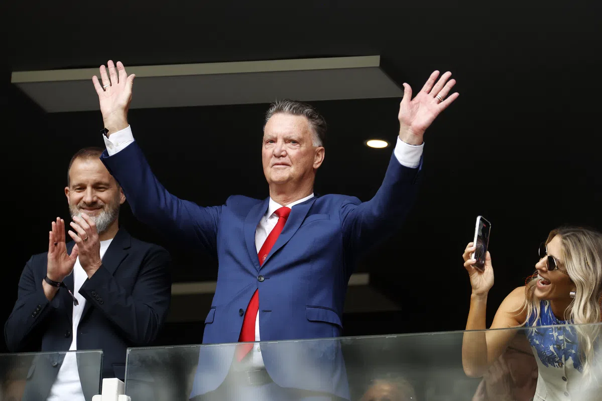 Louis van Gaal staat in een skybox met beide handen omhoog terwijl omstanders applaudisseren en filmen.