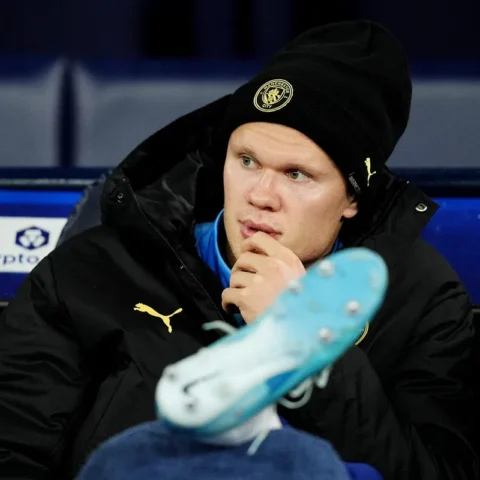 Erling Haaland zit op de bank in een Manchester City-muts en jas tijdens een wedstrijd.