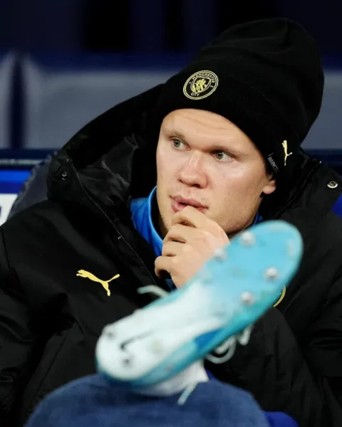 Erling Haaland zit op de bank in een Manchester City-muts en jas tijdens een wedstrijd.
