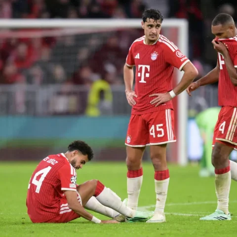Bayern München-spelers op het veld tijdens een Champions League-wedstrijd, met een speler zittend op de grond en twee teamgenoten naast hem.