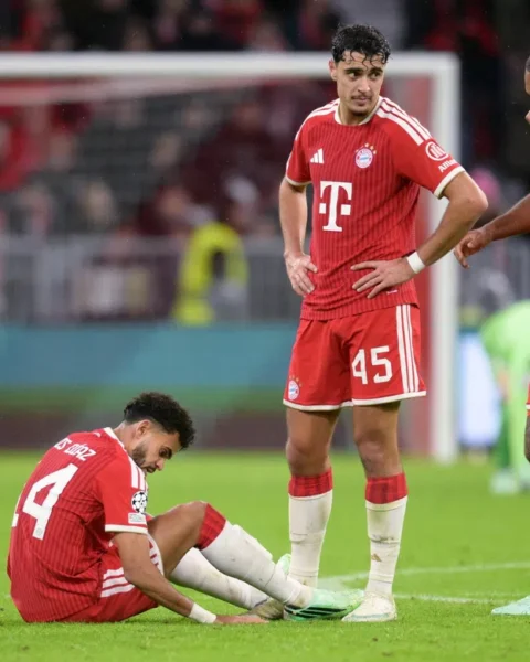 Bayern München-spelers op het veld tijdens een Champions League-wedstrijd, met een speler zittend op de grond en twee teamgenoten naast hem.