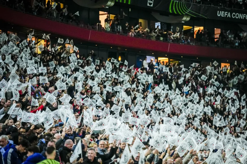 Volgepakte tribunes in een voetbalstadion met veel wapperende witte vlaggen tijdens een Conference League-wedstrijd.