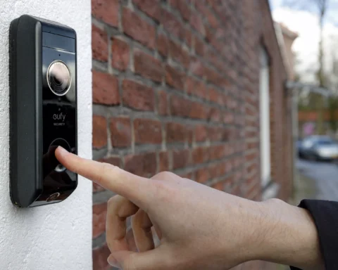 Een hand drukt op een slimme deurbel met camera naast een huisgevel.
