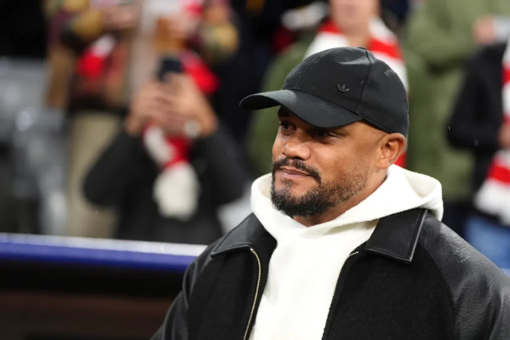 Vincent Kompany draagt een zwarte pet en witte hoodie bij een voetbalwedstrijd.