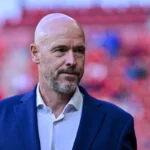Erik ten Hag staat in een stadion voor een wazige rode achtergrond.