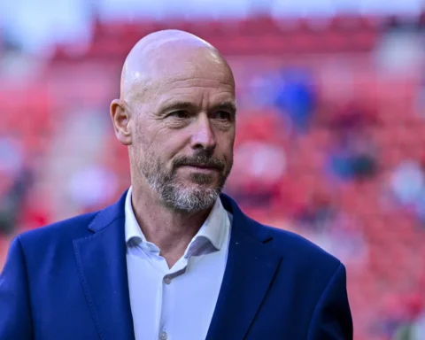 Erik ten Hag staat in een stadion voor een wazige rode achtergrond.
