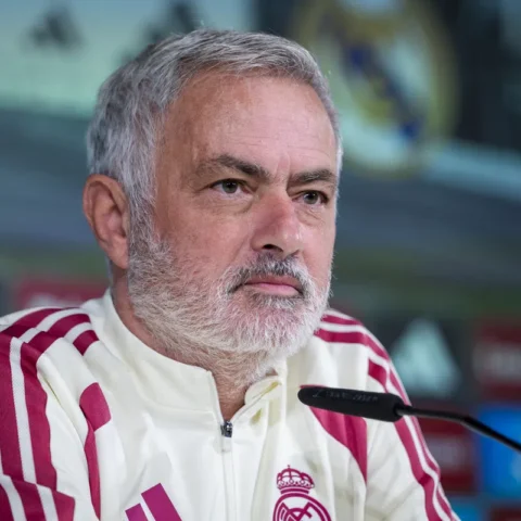 José Mourinho zit aan tafel tijdens een persmoment in een wit trainingsjack.