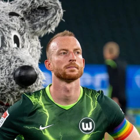 Maximilian Arnold in een VfL Wolfsburg-shirt met regenboogband, met de Wolfsburg-mascotte op de achtergrond.