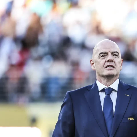 Gianni Infantino staat in een blauw pak met FIFA-logo op de borst tijdens een evenement op een voetbalveld.