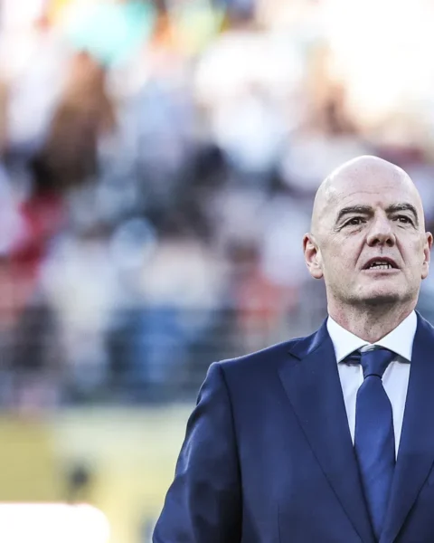 Gianni Infantino staat in een blauw pak met FIFA-logo op de borst tijdens een evenement op een voetbalveld.