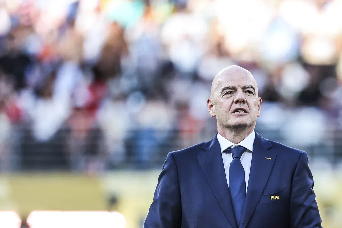 Gianni Infantino staat in een blauw pak met FIFA-logo op de borst tijdens een evenement op een voetbalveld.