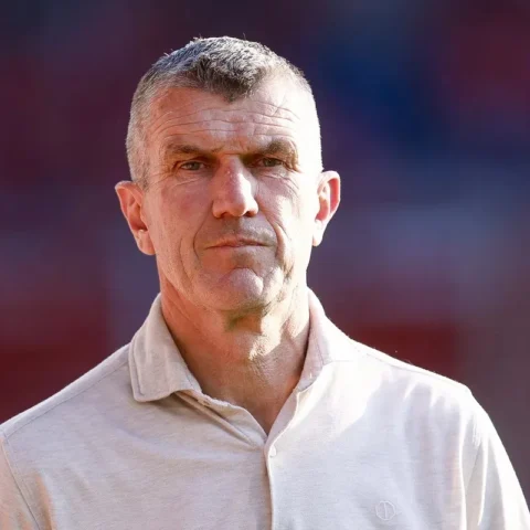 Portret van een man in een licht polohemd op een stadionachtergrond.