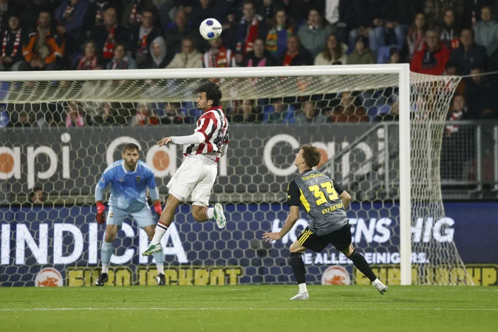 Speler van Willem II springt voor het doel op een voetbalveld terwijl de bal in de lucht hangt.