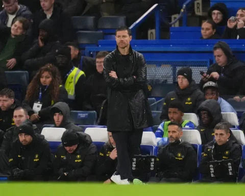 Xabi Alonso staat met gekruiste armen langs de zijlijn tijdens een voetbalwedstrijd, met Chelsea-spelers en staf op de bank achter hem.