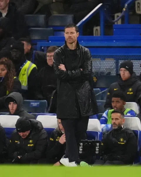 Xabi Alonso staat met gekruiste armen langs de zijlijn tijdens een voetbalwedstrijd, met Chelsea-spelers en staf op de bank achter hem.