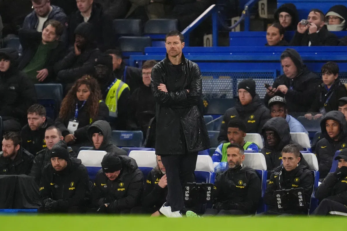Xabi Alonso staat met gekruiste armen langs de zijlijn tijdens een voetbalwedstrijd, met Chelsea-spelers en staf op de bank achter hem.