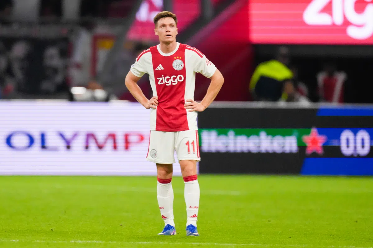 Ajax-speler in wit-rood tenue staat op het veld met handen in de zij.