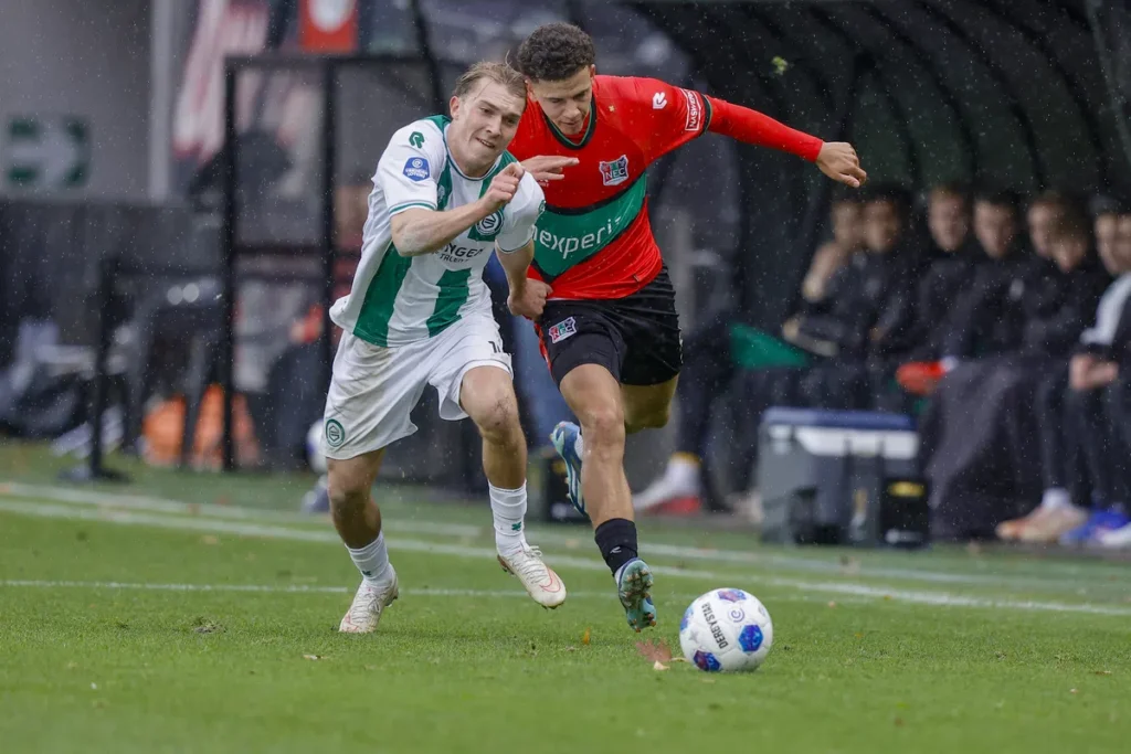 Twee voetballers sprinten achter de bal aan in de regen tijdens NEC tegen FC Groningen.