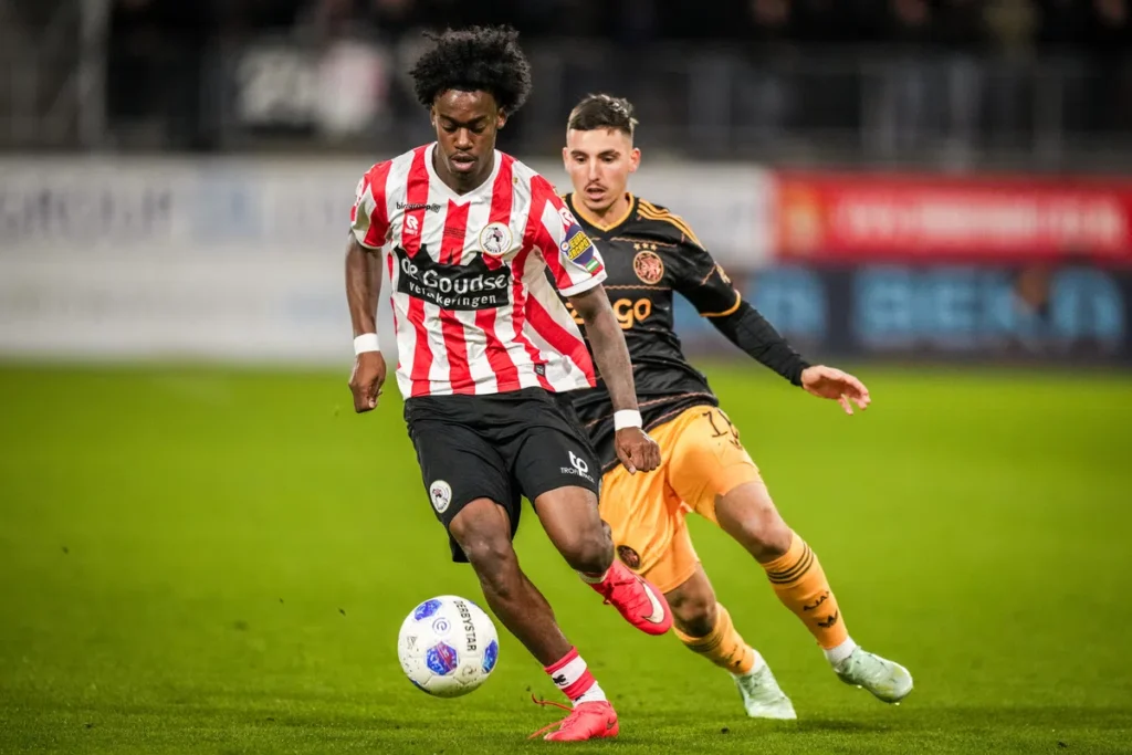 Speler van Sparta Rotterdam in een rood-wit gestreept shirt dribbelt de bal terwijl een Ajax-speler hem op de voet volgt.