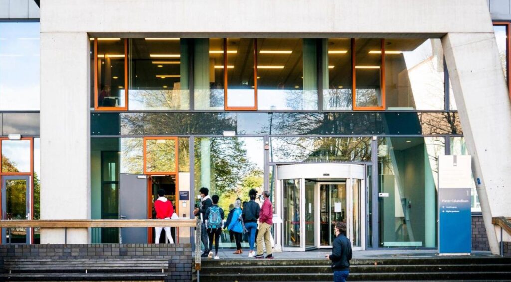 Mensen staan bij de ingang van een modern overheidsgebouw met draaideur en glazen gevel.
