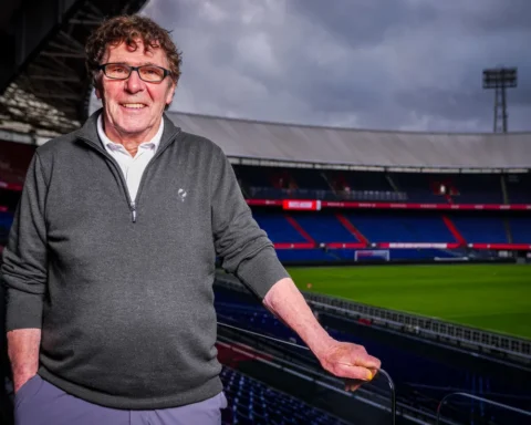 Willem van Hanegem staat in een voetbalstadion met het veld en de tribunes op de achtergrond.