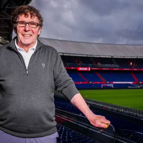 Willem van Hanegem staat in een voetbalstadion met het veld en de tribunes op de achtergrond.