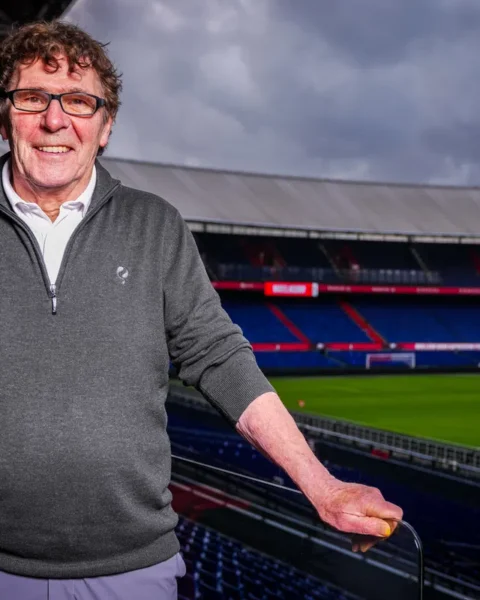Willem van Hanegem staat in een voetbalstadion met het veld en de tribunes op de achtergrond.