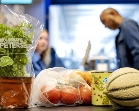 Supermarkttoonbank met biologische peterselie, groenten en andere boodschappen; op de achtergrond staan twee winkelmedewerkers.