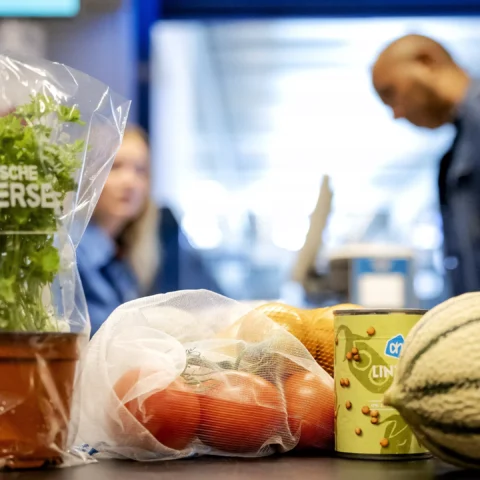 Supermarkttoonbank met biologische peterselie, groenten en andere boodschappen; op de achtergrond staan twee winkelmedewerkers.