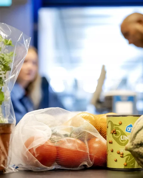 Supermarkttoonbank met biologische peterselie, groenten en andere boodschappen; op de achtergrond staan twee winkelmedewerkers.