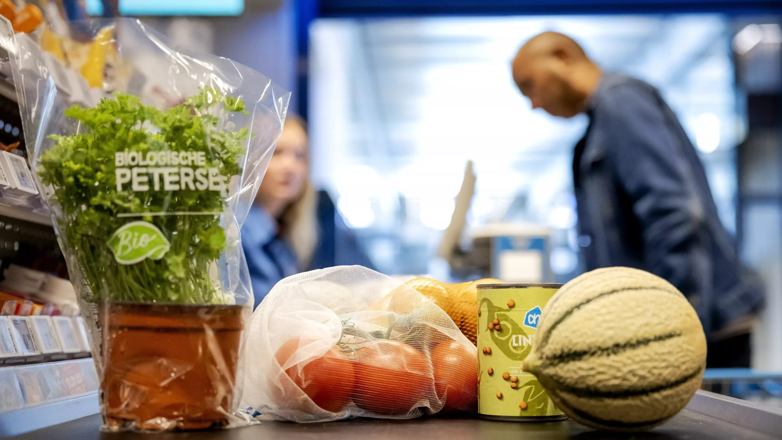 Supermarkttoonbank met biologische peterselie, groenten en andere boodschappen; op de achtergrond staan twee winkelmedewerkers.
