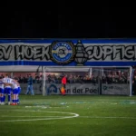 Voetbalteam in een huddle op het veld voor een goal, met supporters achter het doel.