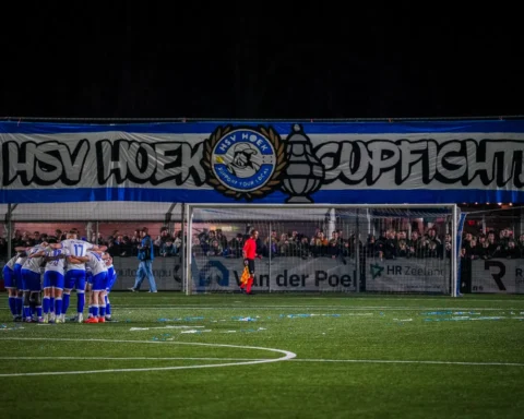 Voetbalteam in een huddle op het veld voor een goal, met supporters achter het doel.