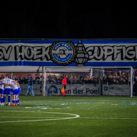 Voetbalteam in een huddle op het veld voor een goal, met supporters achter het doel.