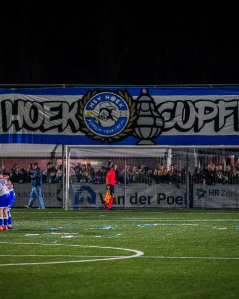 Voetbalteam in een huddle op het veld voor een goal, met supporters achter het doel.