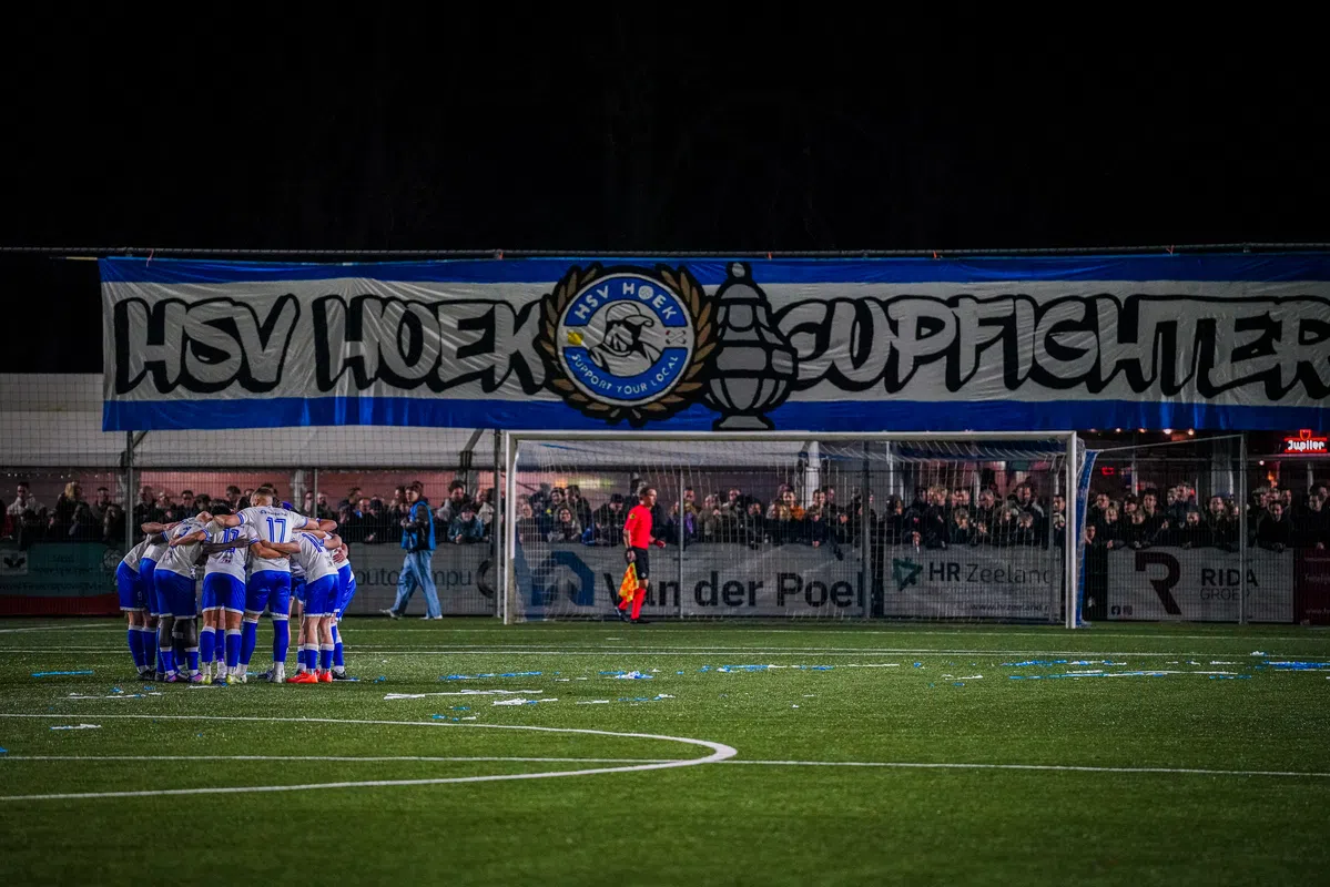 Voetbalteam in een huddle op het veld voor een goal, met supporters achter het doel.