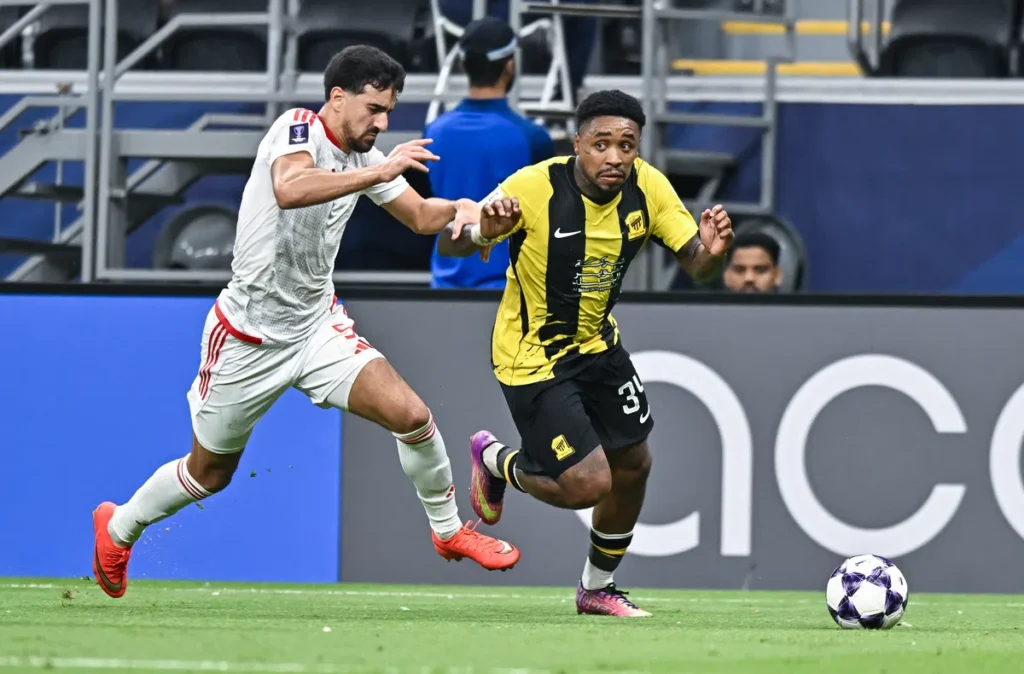 Twee voetballers sprinten om de bal tijdens een wedstrijd, met Al-Ittihad in geel-zwart.