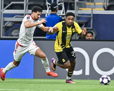 Twee voetballers sprinten om de bal tijdens een wedstrijd, met Al-Ittihad in geel-zwart.