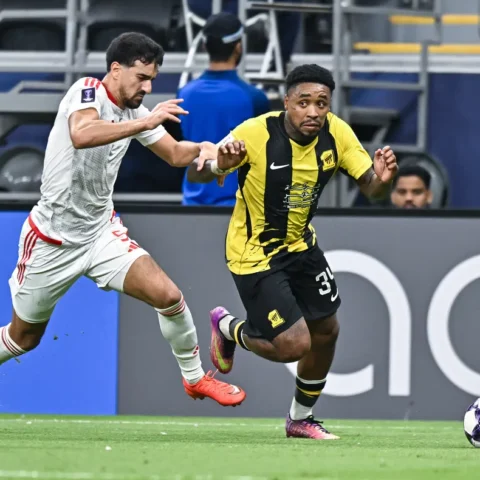 Twee voetballers sprinten om de bal tijdens een wedstrijd, met Al-Ittihad in geel-zwart.