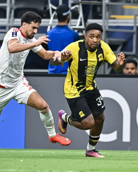 Twee voetballers sprinten om de bal tijdens een wedstrijd, met Al-Ittihad in geel-zwart.