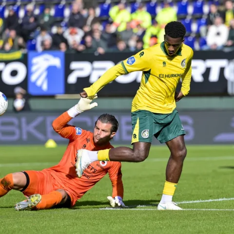 Speler in geel-groen en doelman in oranje tijdens een voetbalduel in het strafschopgebied.