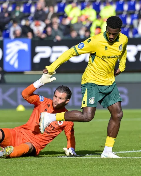 Speler in geel-groen en doelman in oranje tijdens een voetbalduel in het strafschopgebied.