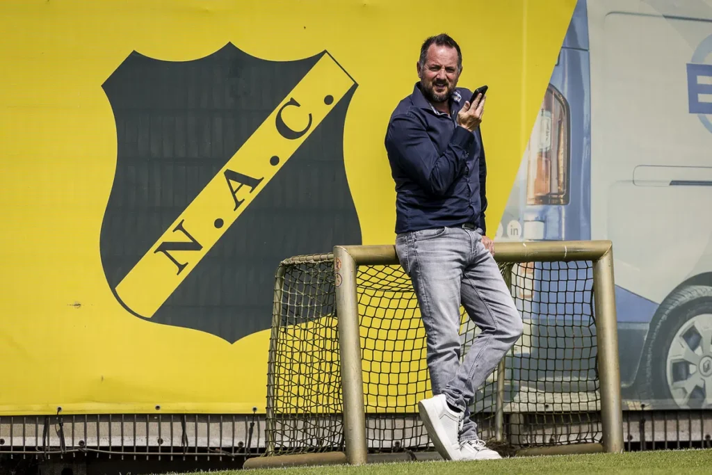 Man staat voor een groot NAC-logo bij een voetbaltrainingscomplex.
