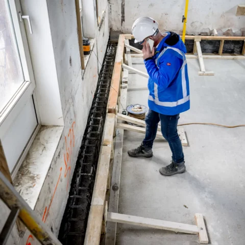 Een bouwmedewerker in beschermende kleding inspecteert funderingsherstel in een gebouw.