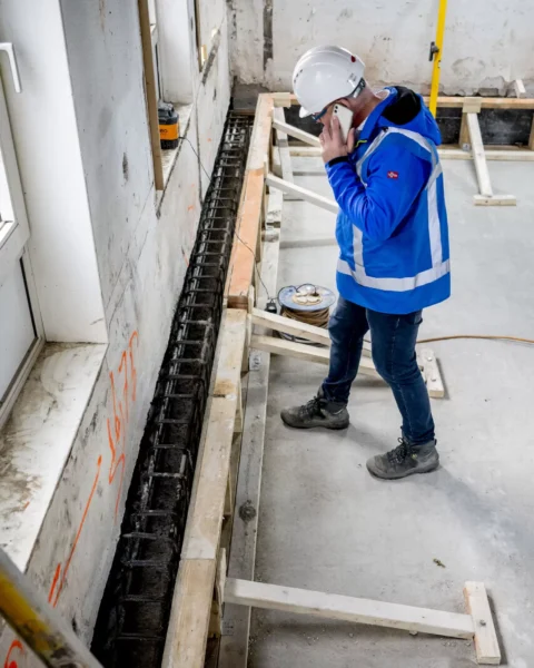 Een bouwmedewerker in beschermende kleding inspecteert funderingsherstel in een gebouw.