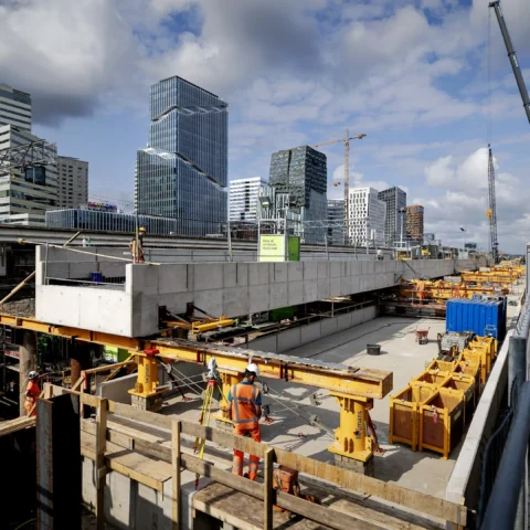 Bouwplaats in een moderne skyline met kranen, werknemers en voorbijlopende mensen op een loopbrug.