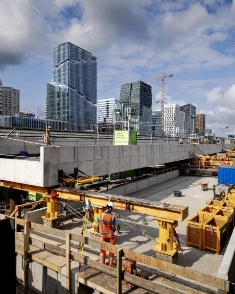 Bouwplaats in een moderne skyline met kranen, werknemers en voorbijlopende mensen op een loopbrug.